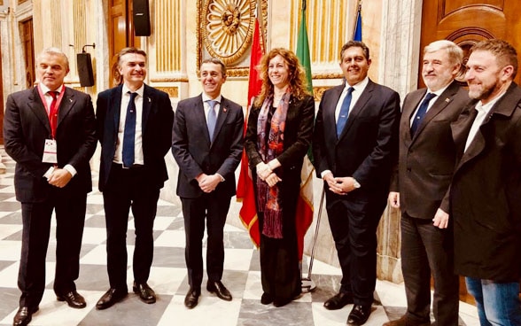 Le conseiller fédéral Ignazio Cassis (troisième à partir de la gauche), une femme et cinq autres hommes se tiennent en demi-cercle devant trois drapeaux.