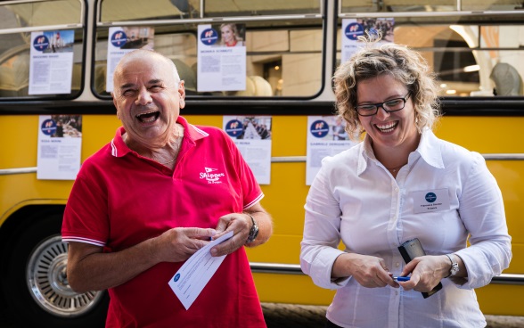 A visitor in a red shirt laughs with an ambassador; in the background is the bus Meet the Ambassador