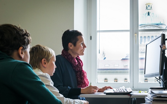 Two children watch a woman work at a computer.