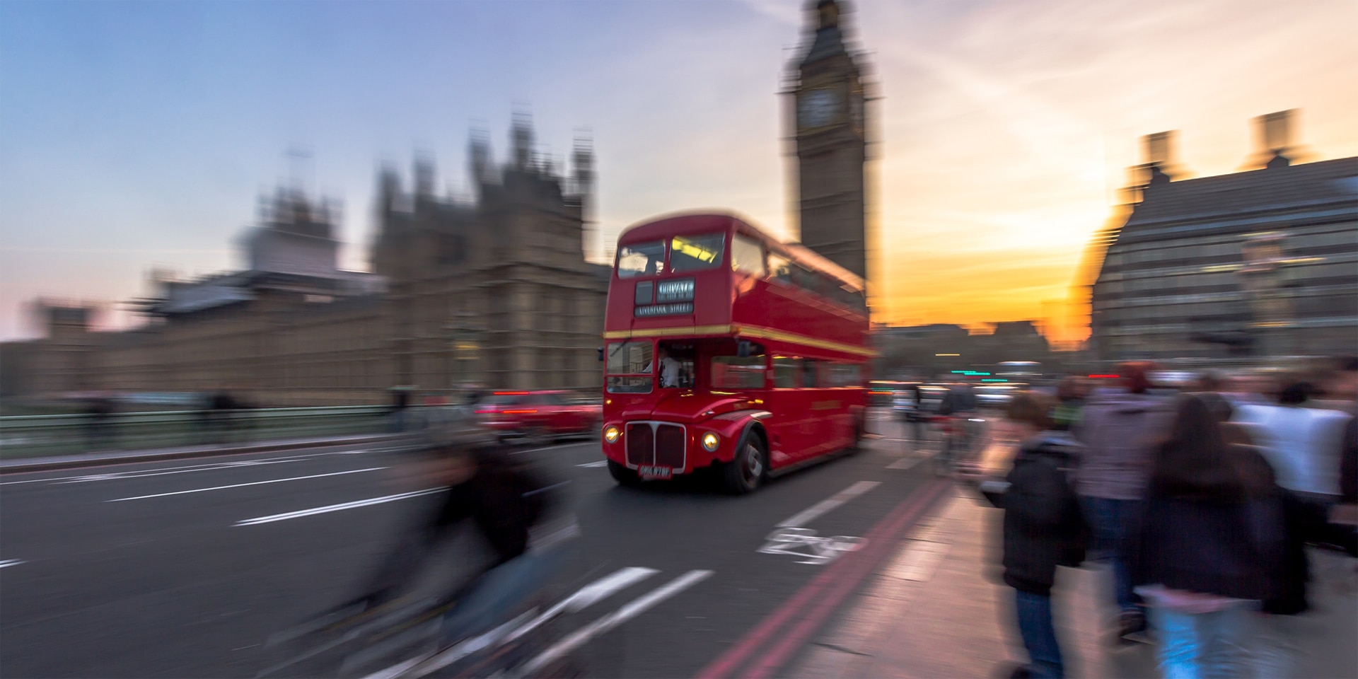 Ein für London typischer roter Doppeldeckerbus fährt vorbei am Big Ben über die Westminster Bridge ins Abendrot. 