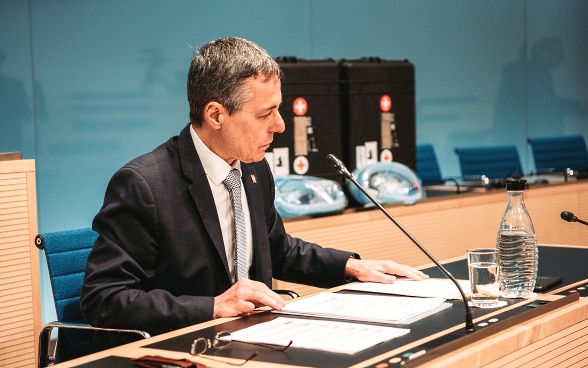 Bundesrat Cassis sitzt an einem Pult, Dossiers liegen vor ihm auf dem Tisch. Im Hintergrund sieht man die beiden Koffer mit den Beatmungsgeräten.