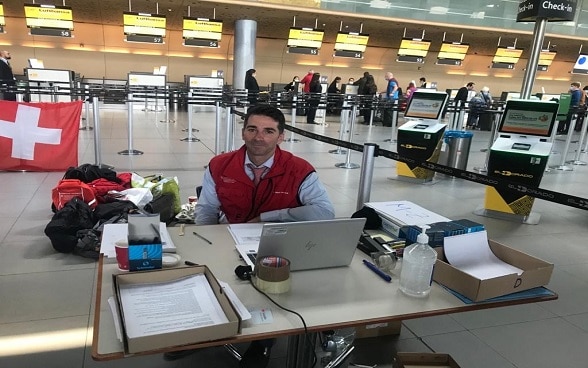 In der Check-in-Halle des Flughafens Bogotá sitzt ein Mitglied der Schweizer Botschaft in Kolumbien hinter einem Tisch und einem Computer, um Reisenden zu helfen 