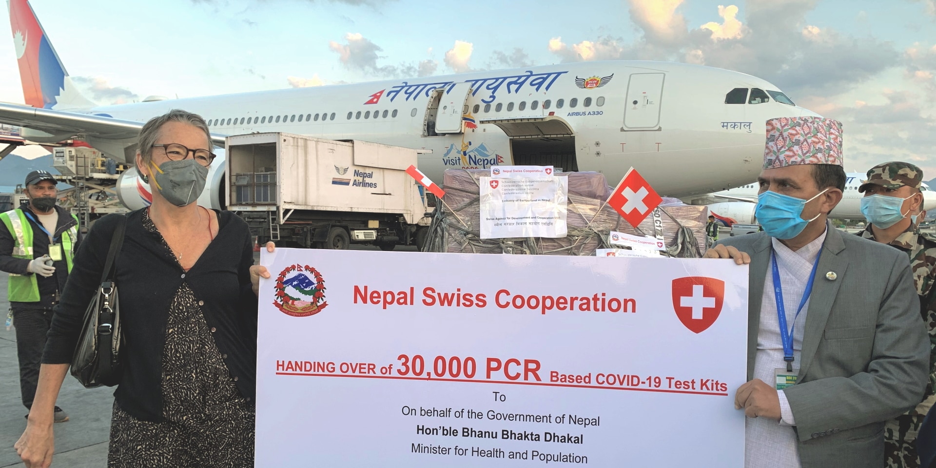 Die Schweizer Botschafterin in Nepal und Nepals Minister für Gesundheit und Bevölkerung stehen vor einem Flugzeug und halten ein Plakat in der Hand, in der die Übergabe von 30'000 COVID-19-Testkits angezeigt wird.
