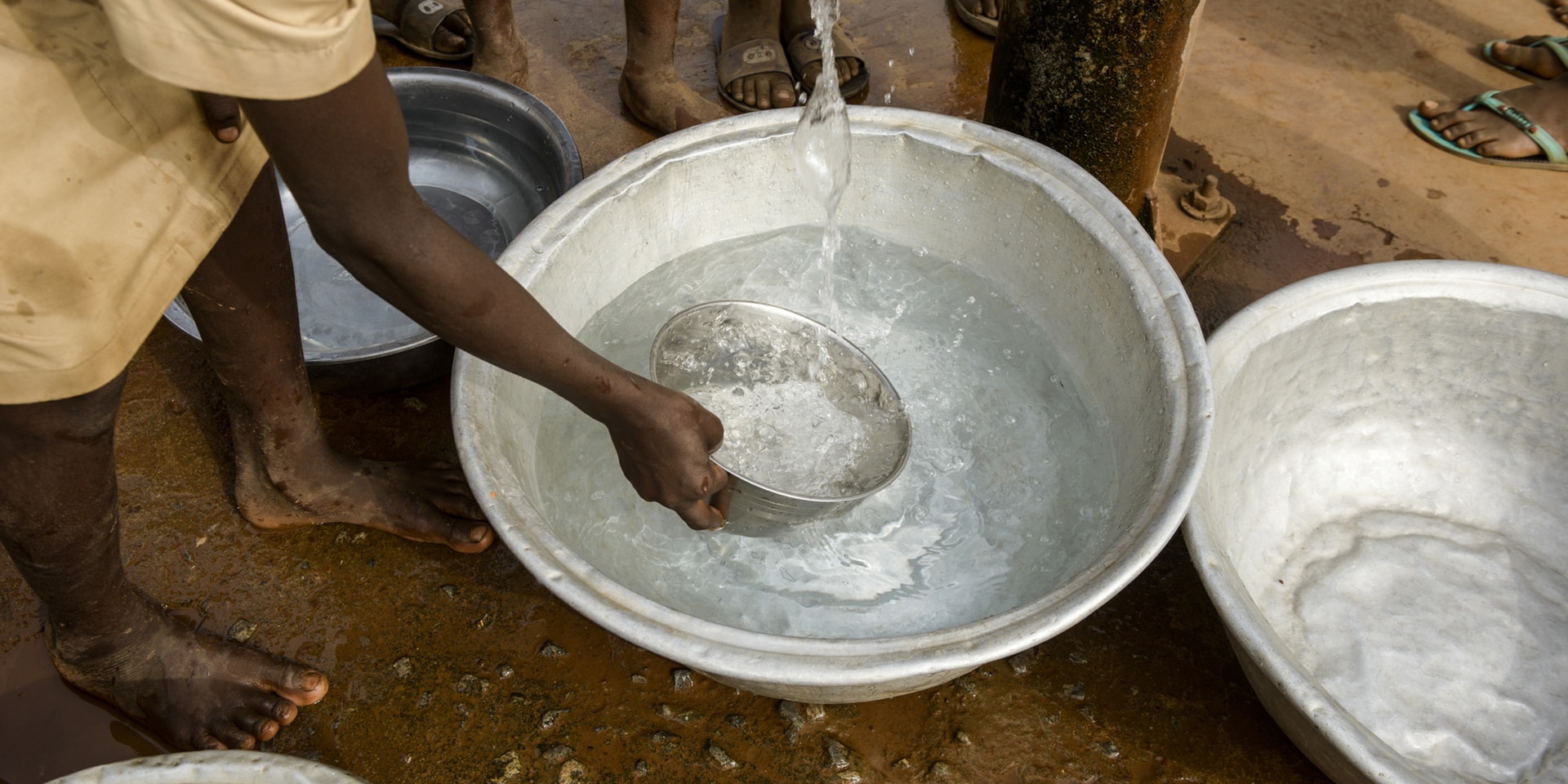 Mehrere Personen stehen an einem Brunnen und schöpfen mit einer Schale sauberes Wasser. 