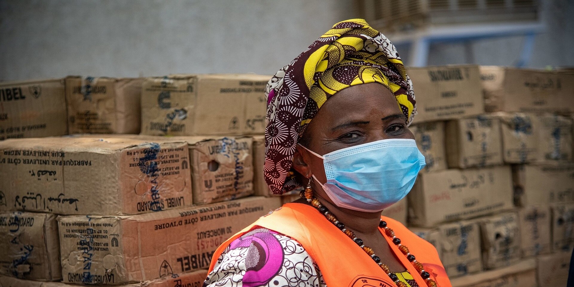 Una donna africana con una mascherina e un gilet arancione lavora in un deposito dove sono immagazzinate delle scatole.