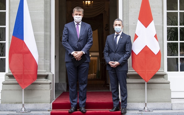 Ignazio Cassis und Tomáš Petříček stehen vor dem Eingang eines Gebäudes, neben ihnen die Flaggen ihrer jeweiligen Länder.