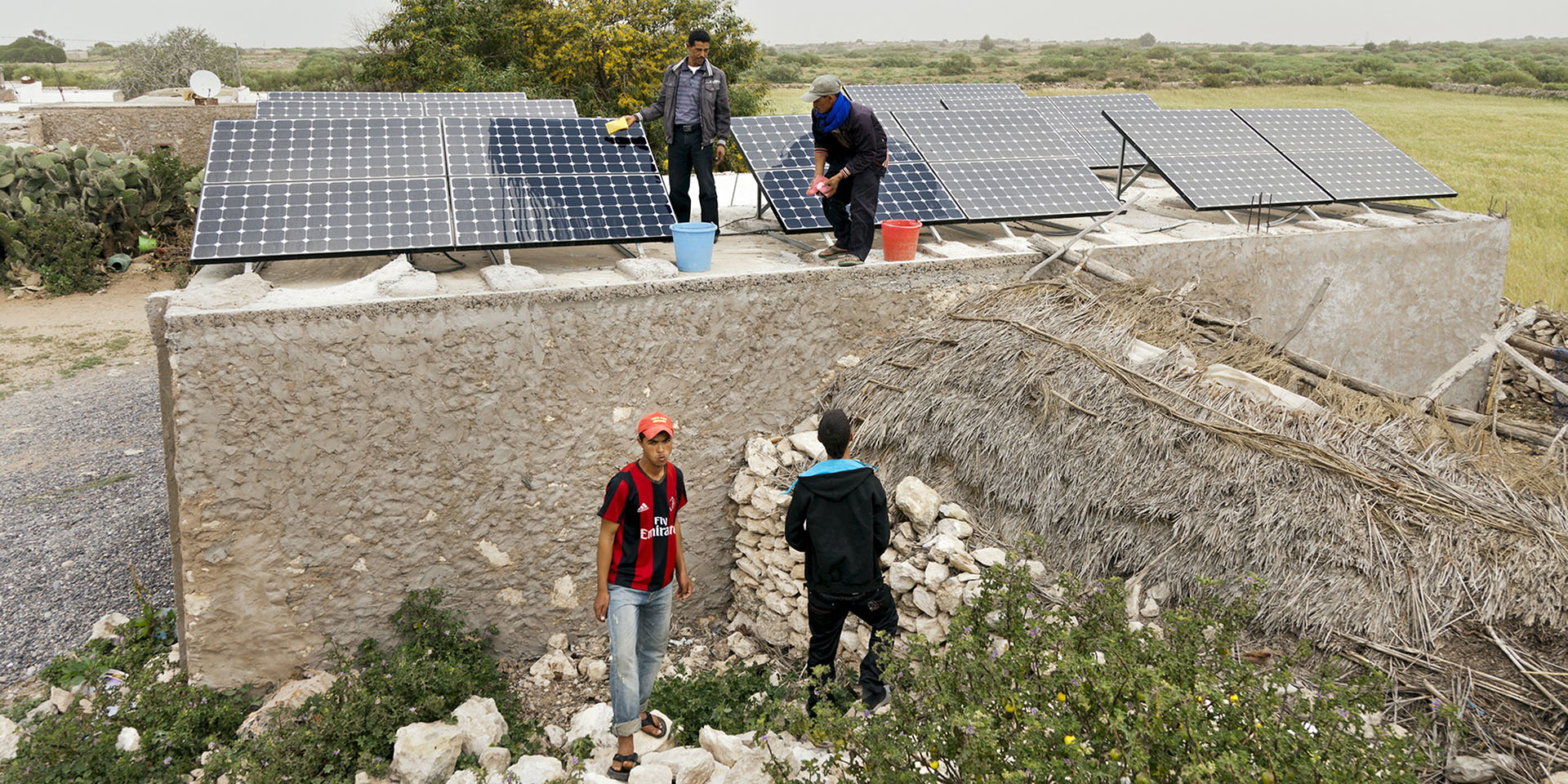 Zwei Männer stehen auf einem Steingebäude und waschen Solarpanels. Neben dem Haus stehen zwei weitere Männer.