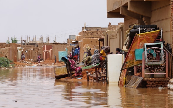 Alcune persone sostano ai bordi della strada sommersa dall’acqua. Accanto a loro alcuni mobili recuperati.