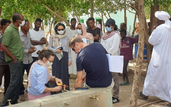Deux experts de l'Aide humanitaire prélèvent des échantillons d'eau. Quelques personnes se tiennent autour d'eux.