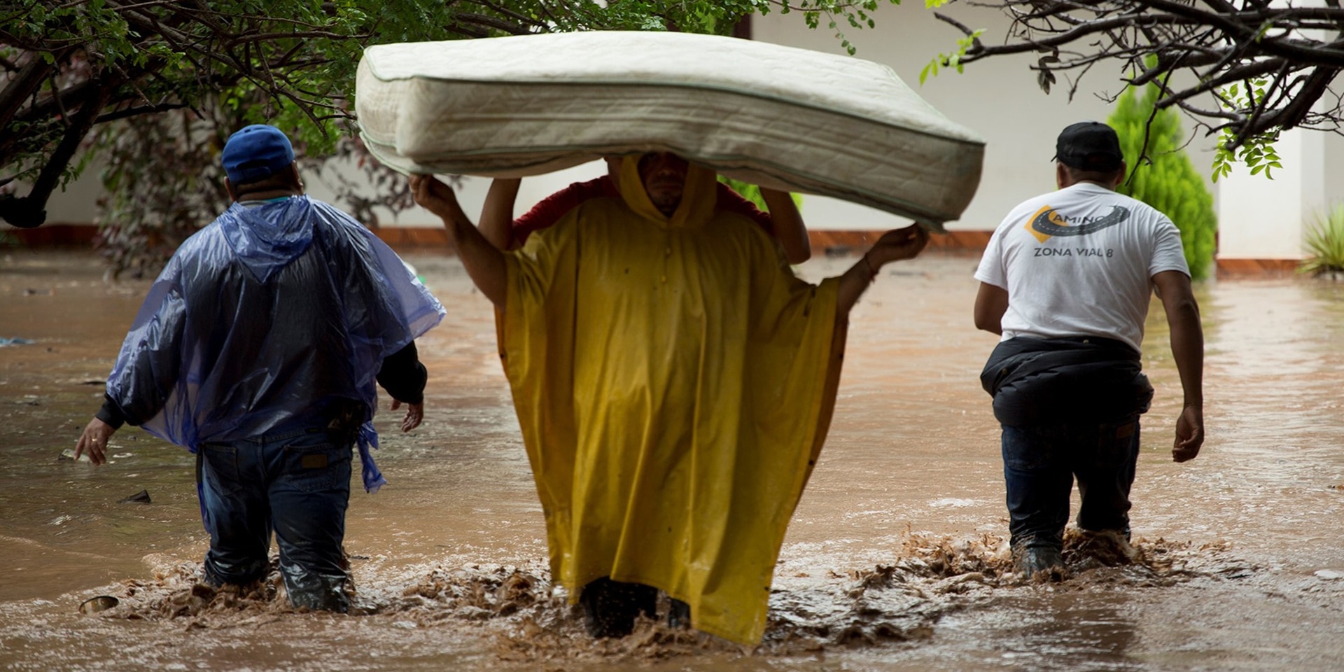 Zwei Personen tragen eine Matratze durch das Hochwasser. Zwei Mitglieder der Hilfskräfte begleiten sie dabei.
