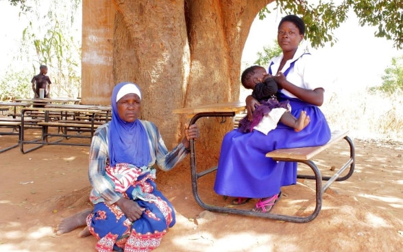 Une femme est assise par terre, à l’ombre d’un arbre. À ses côtés, une autre femme, assise sur une chaise d’école, berce un bébé dans ses bras.