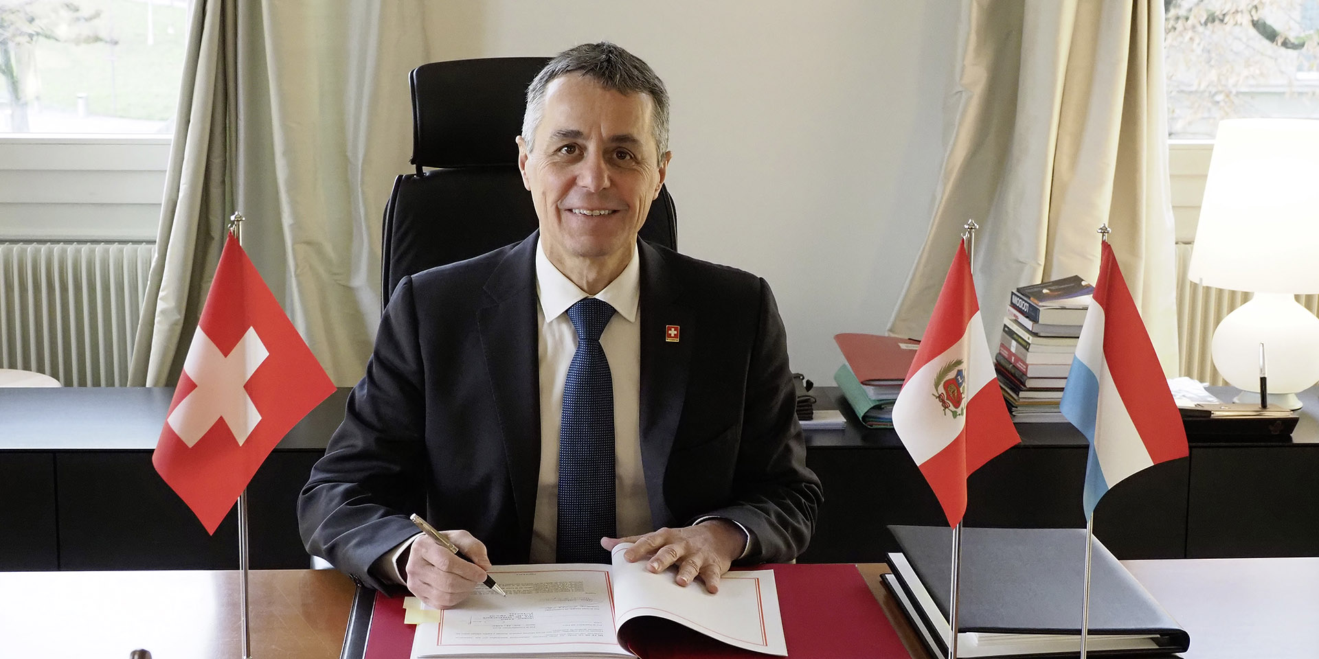 Le Conseiller fédéral Ignazio Cassis signe un document. Sur son bureau, apparaissent les drapeaux de la Suisse (à sa droite), du Pérou et du Luxembourg (à sa gauche).