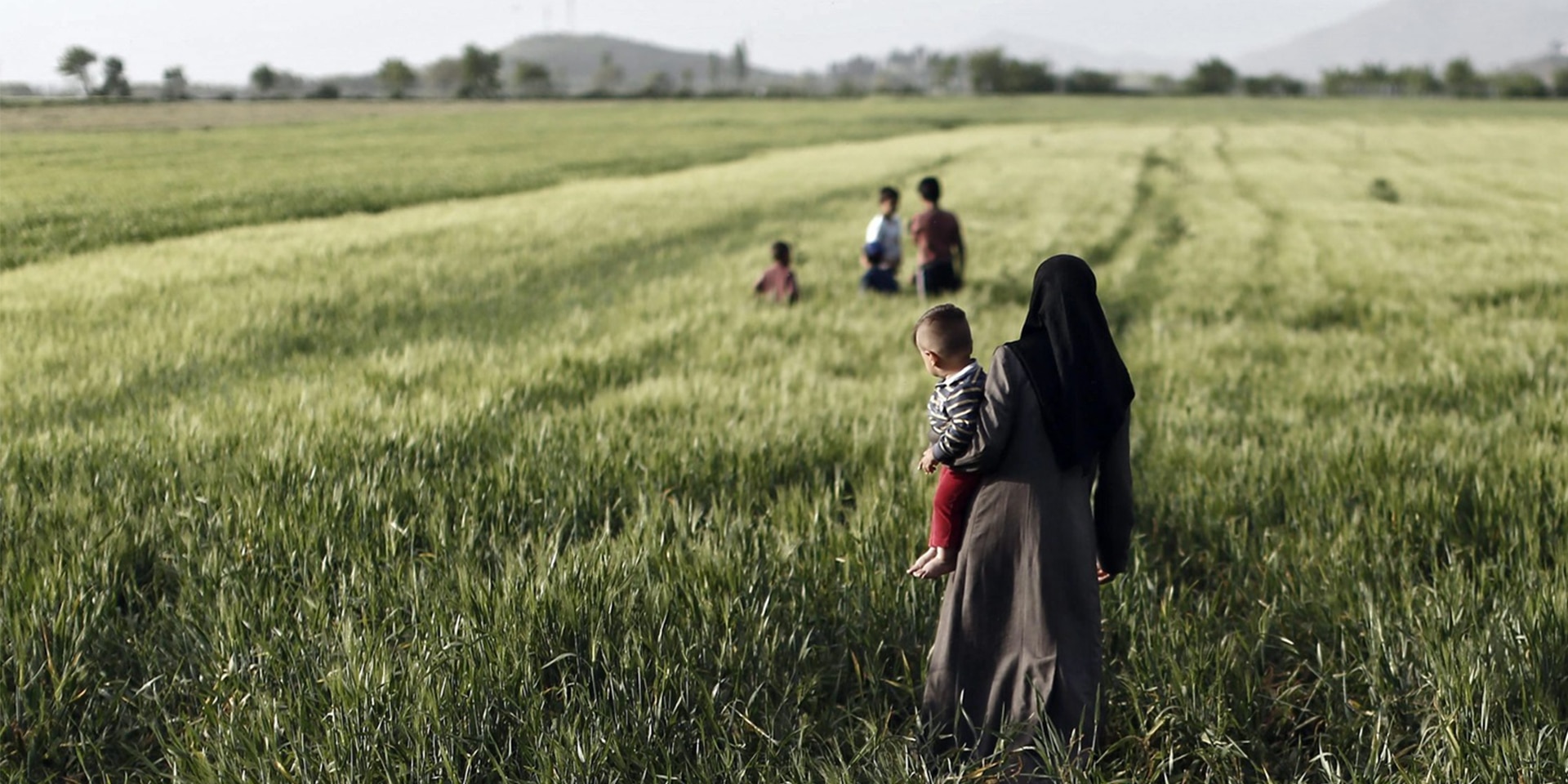 A woman with an infant in her arms stands in a field with high grass and watches children playing in the distance.