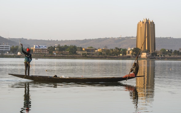 Zwei Fischer auf einem Holzboot fischen vor der Skyline Bamakos.