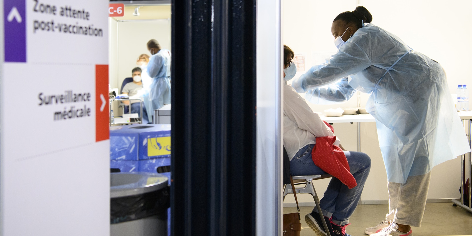 Dans un centre de vaccination, une infirmière administre un vaccin à un patient. Des panneaux indiquent où se rendre pour les contrôles médicaux et où attendre après avoir reçu le vaccin.