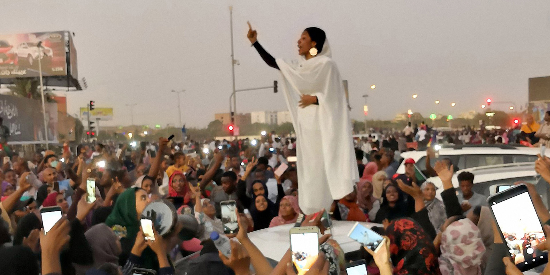 Surrounded by a crowd, a woman standing on the bonnet of a car speaks. She is filmed with numerous mobile phones.   