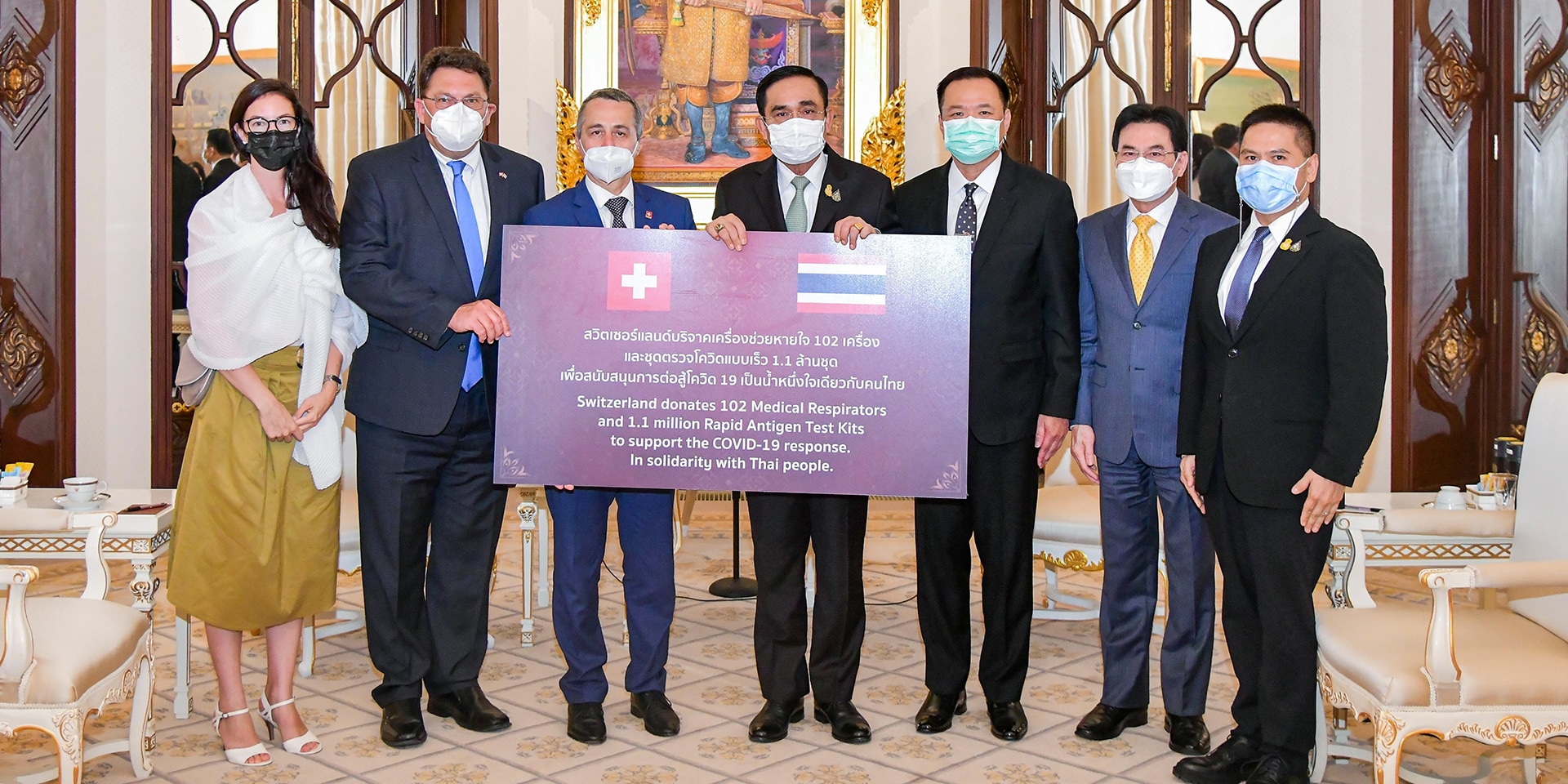 Verschiedene Personen stehen in einem Raum vor einem Schild, welches die Hilfslieferung der Schweiz beschreibt. 