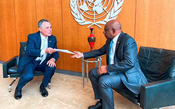 Vor dem Hintergrund des UNO-Emblems überreicht Bundesrat Ignazio Cassis dem Präsidenten der UNO-Generalversammlung Abdulla Shahid ein Dokument (beide sitzend).