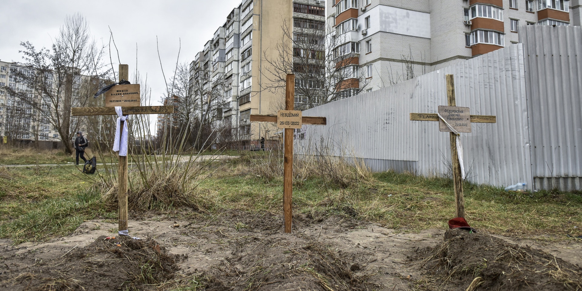 Trois tombes avec des croix en bois et des noms sont visibles au premier plan. De grandes maisons se trouvent à l'arrière-plan.