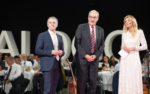 Ignazio Cassis, Guy Parmelin et Christa Rigozzi parlent sur une scène.