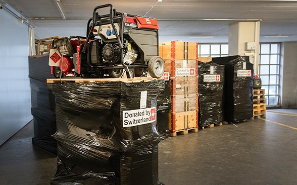  In un magazzino, una scatola con l'immagine di una stufa mobile su un pallet è in primo piano. Sullo sfondo, altri pallet su cui sono posizionati i generatori di corrente imballati.