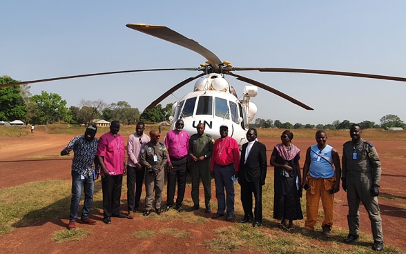 Dieci uomini e una donna africani si trovano davanti a un elicottero bianco delle Nazioni Unite su un terreno sabbioso.