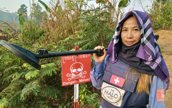 A woman deminer from the HALO Trust wearing an outfit marked with a Swiss cross standing next to a minefield.