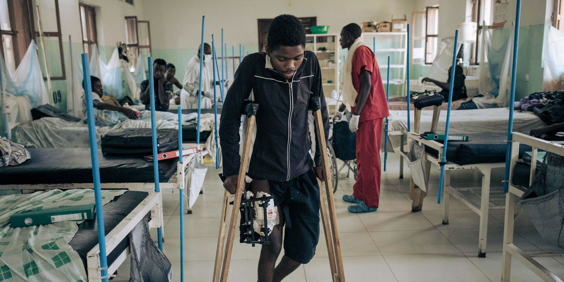 Ein junger afrikanischer Mann mit einer Schusswunde am Bein geht in einem Krankenhaus mühsam auf Krücken.