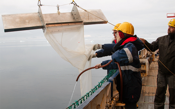 Three scientists handle equipment on a boat and wear yellow helmets on their heads.