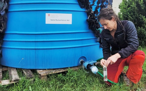 A woman kneels next to a blue water tank and lets water flow into a bottle.