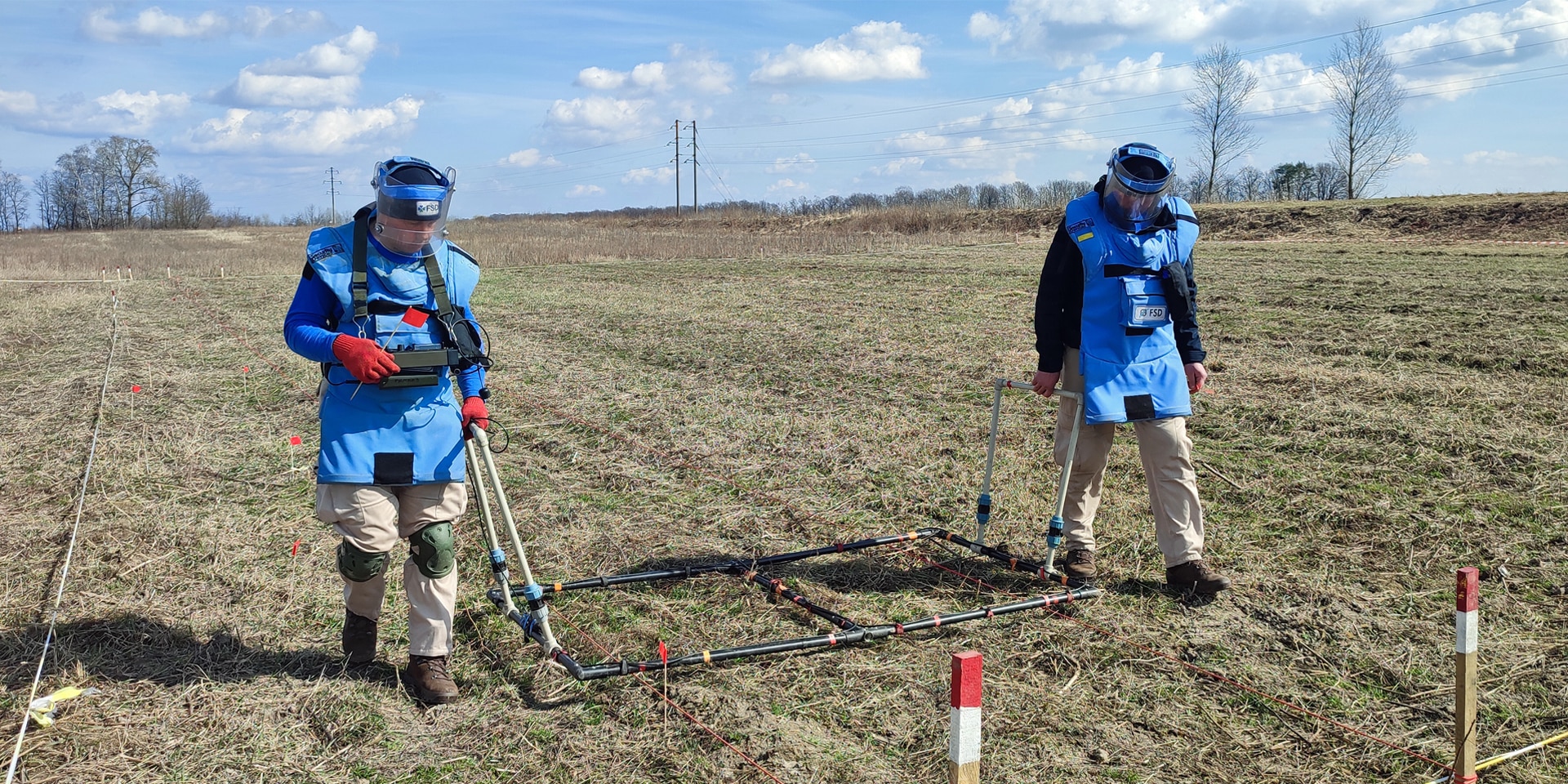 Deux hommes en tenue de protection travaillent au déminage d’un champ où se trouvent probablement des munitions.