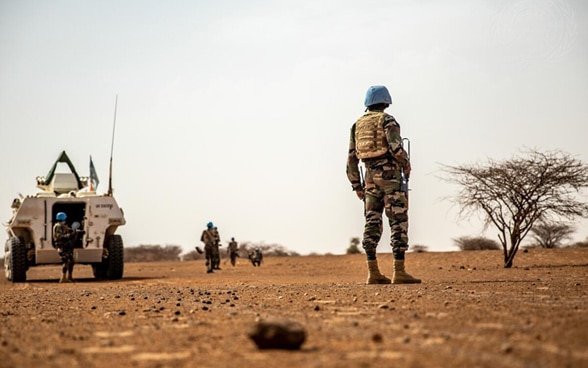 Des casques bleus de la mission de paix de l'ONU (MINUSMA) se tiennent à côté d'un véhicule blindé blanc dans un paysage aride et sablonneux au Mali.