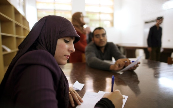 Une jeune femme libyenne portant un foulard violet est assise à une table en bois et prend des notes.