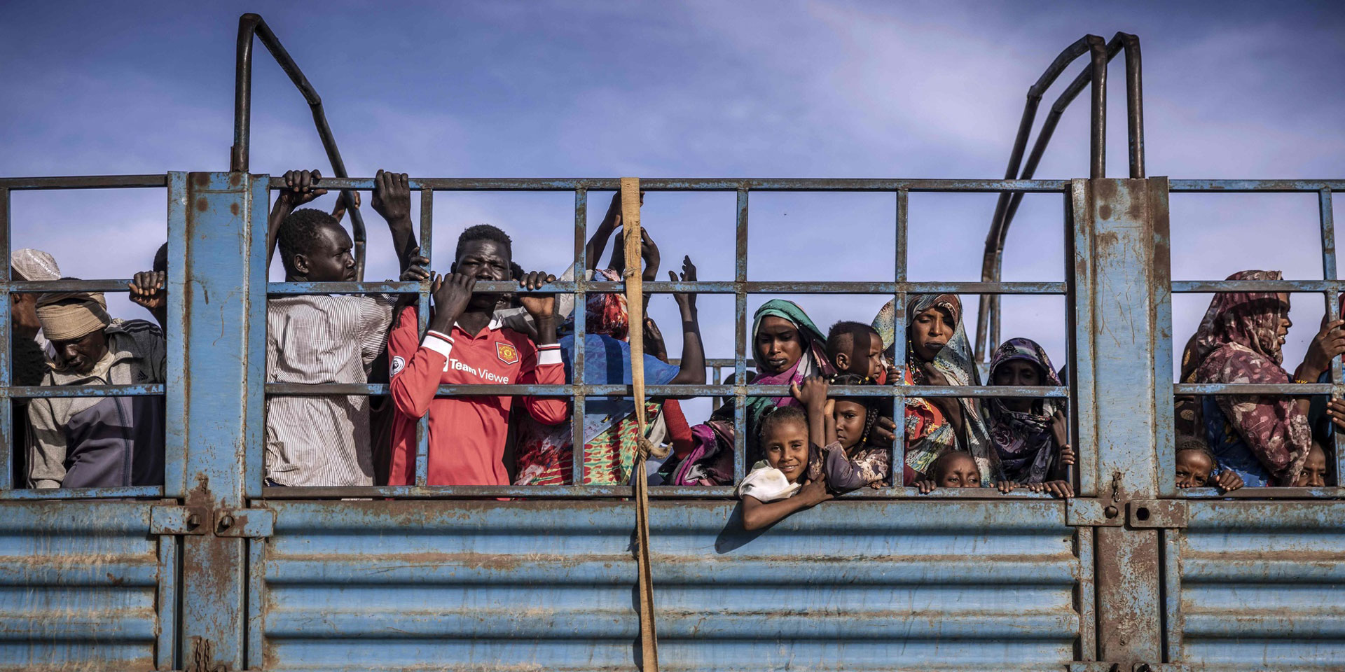 Sudanese women, men and children stand close together on the back of a blue lorry.
