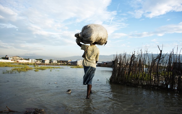 Femme portant un ballot sur sa tête, marchant dans une eau à hauteur de cheville et se dirigeant vers un groupe de maisons. 