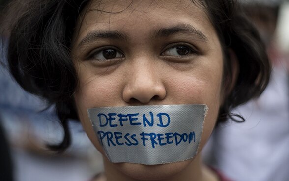 Bambina con la bocca coperta da un pezzo di nastro adesivo su cui campeggia la scritta «defend press freedom».