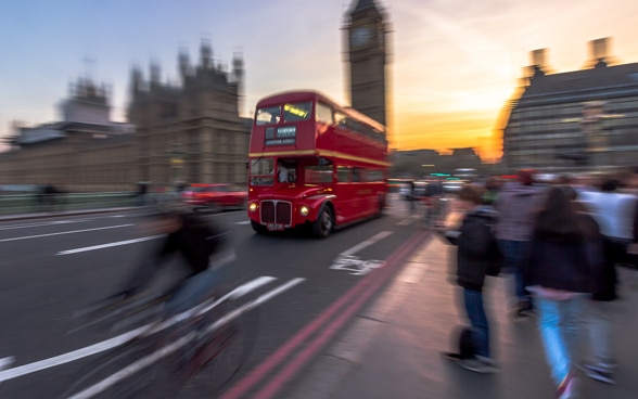 Ein für London typischer roter Doppeldeckerbus fährt vorbei am Big Ben über die Westminster Bridge ins Abendrot. 