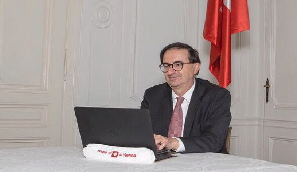 L’ambassadeur Wolfgang Amadeus Brülhart travaille à domicile sur son ordinateur portable. Un drapeau suisse est visible en arrière-plan.