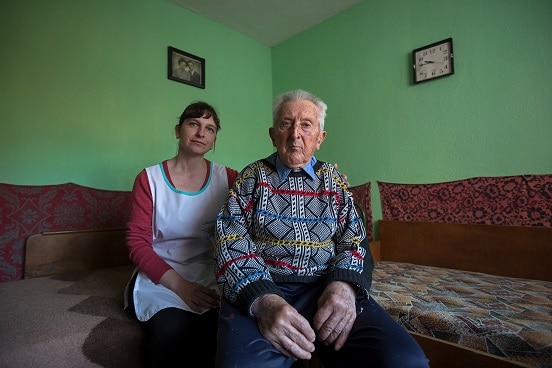 Ein alter Mann sitzt mit einer Pflegeperson auf einem Sofa in seinem Haus.