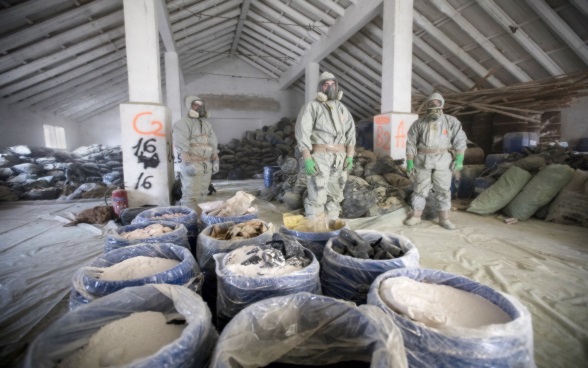 Trois hommes en combinaison de protection derrière des fûts contenant des substances toxiques.