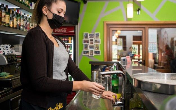 Dietro al bancone di un bar una donna riempie un bicchiere con acqua del rubinetto.