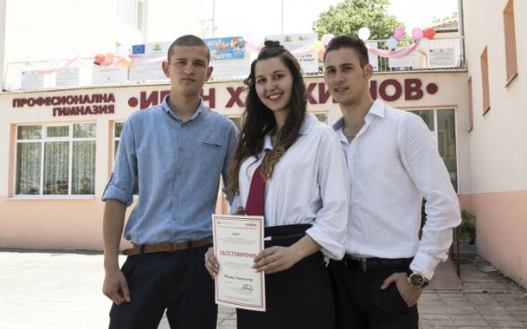 Due ragazzi e una ragazza, che tiene in mano un certificato, in piedi di fronte a una scuola.