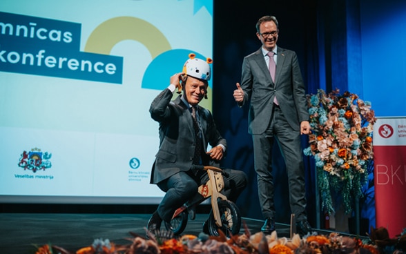 Zwei Männer halten eine Präsentation zum Jubiläum des Spitals, einer sitzt auf einem Kinderfahrrad.