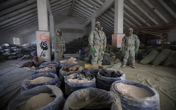 Three men in protective suits in front of several containers with toxic substances