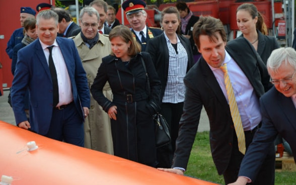 A group of men and women in suits and uniforms look at an orange flood protection barrier.